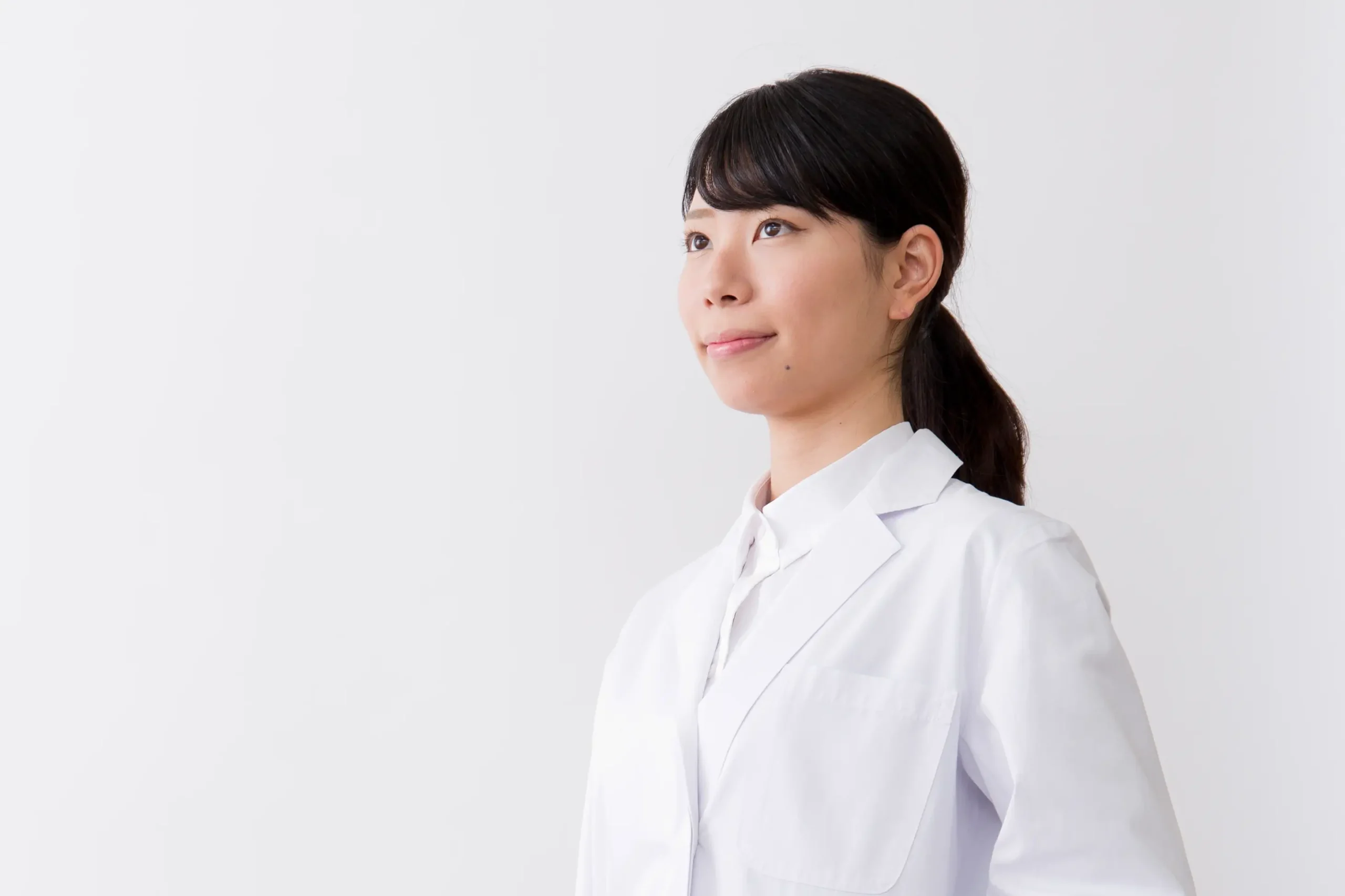 A person with long dark hair tied back, wearing a white lab coat, stands against a plain white background. They are looking slightly upward with a thoughtful expression.