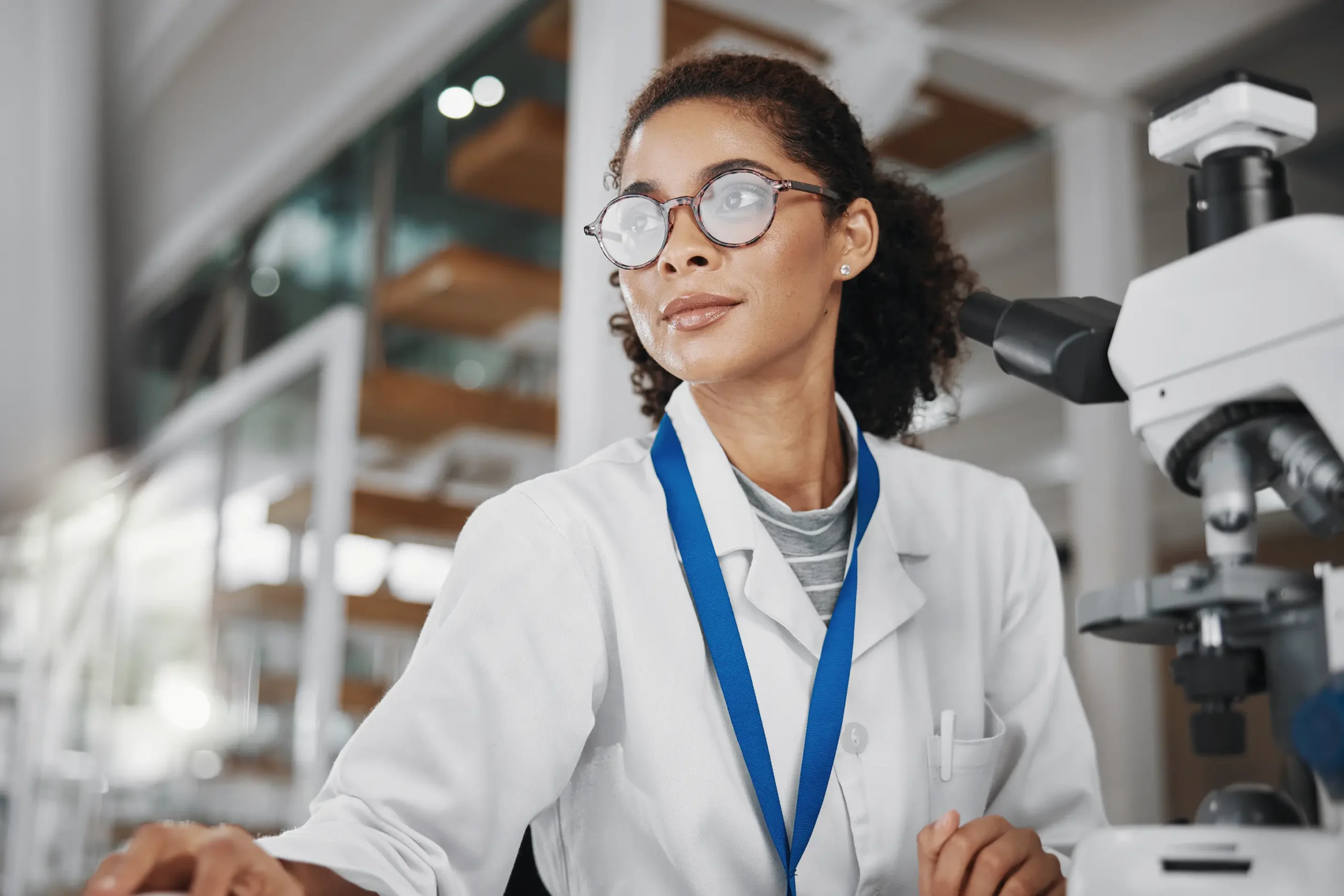 A scientist in a lab coat sits at a desk, looking towards a computer screen. She wears glasses and a blue lanyard. A microscope is visible nearby, and there are shelves and large windows in the background.