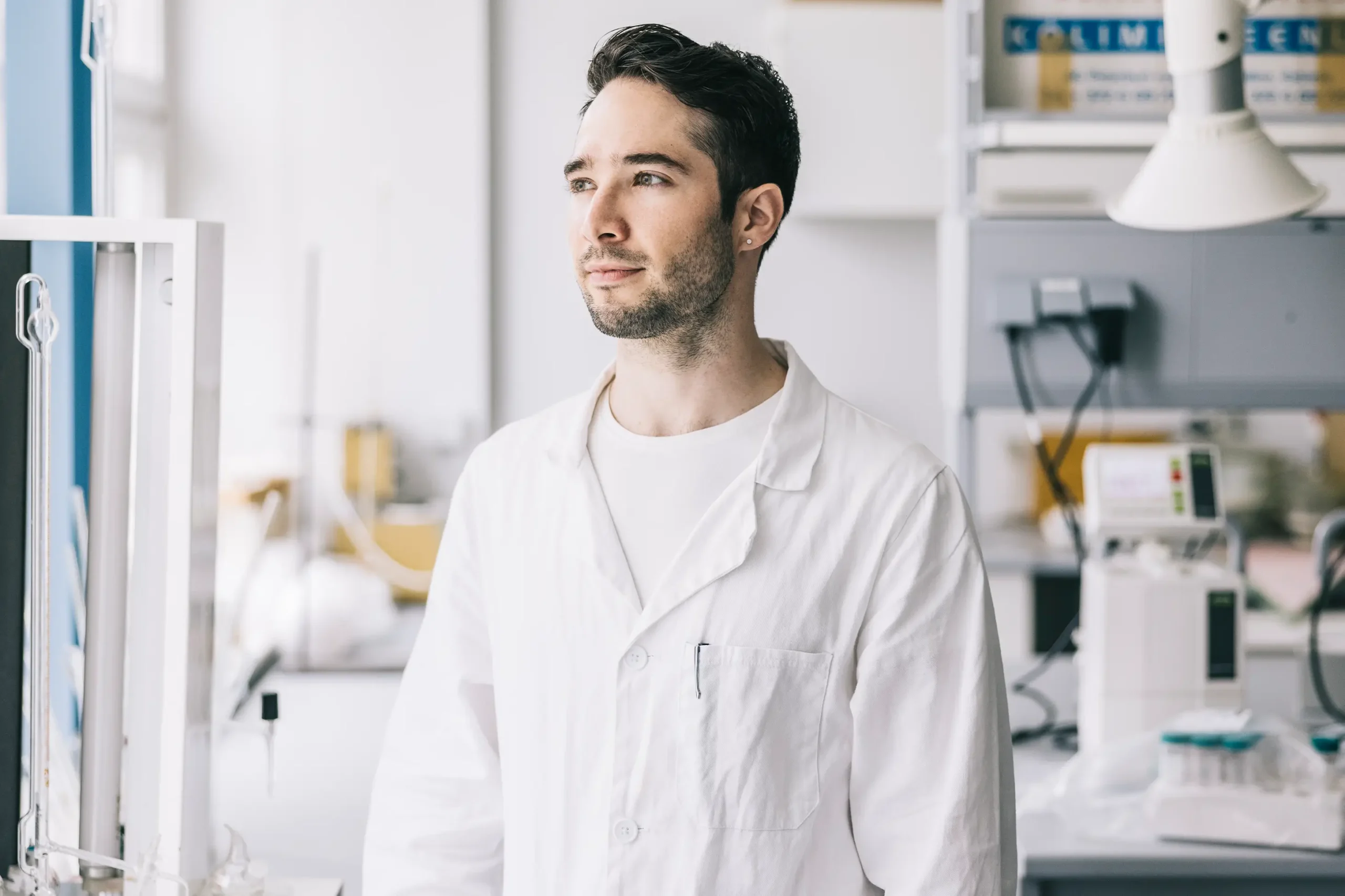 A man in a white lab coat stands in a laboratory, looking thoughtfully into the distance. The lab is filled with scientific equipment and shelves, creating a bright and professional environment.