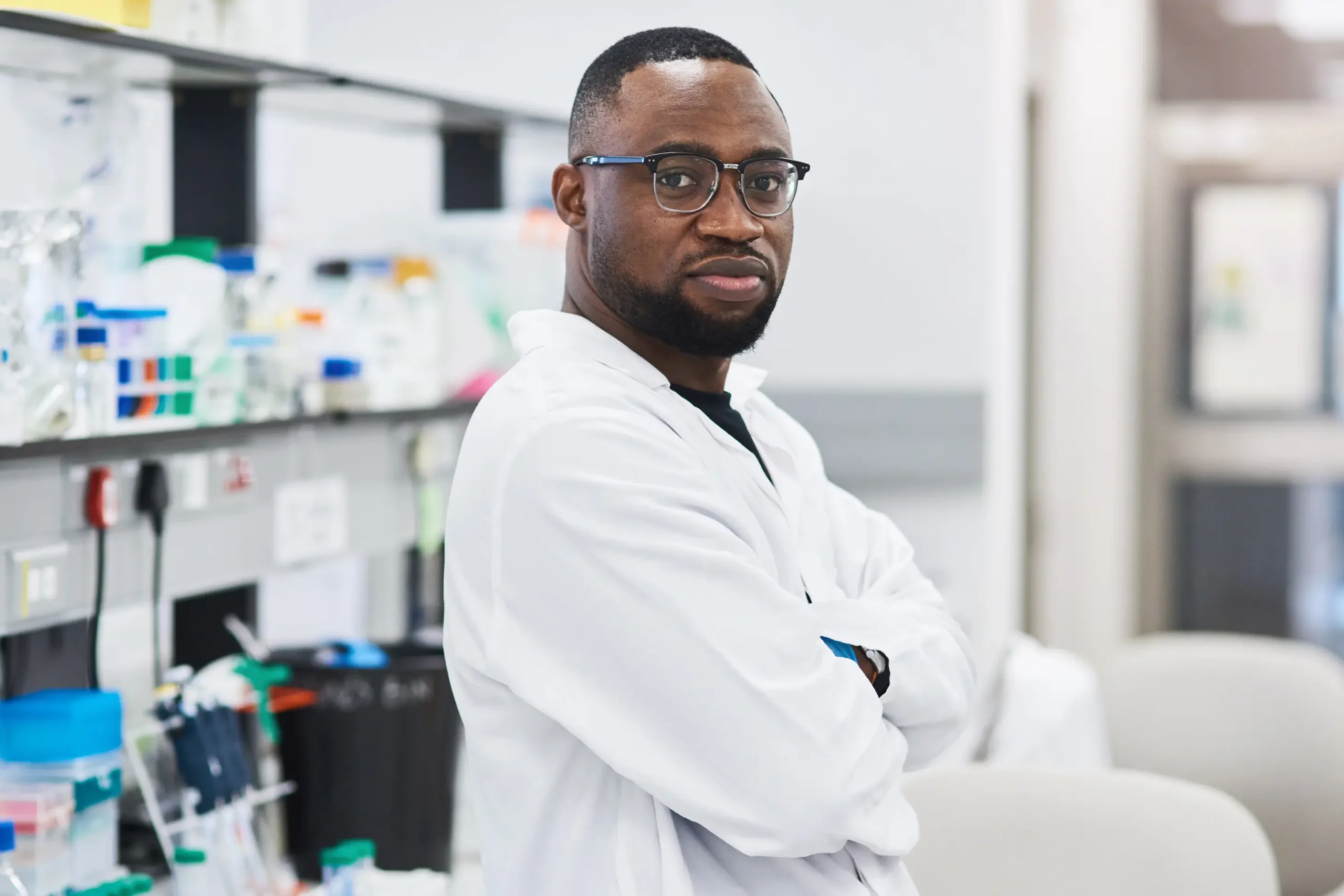 A scientist wearing glasses and a white lab coat stands with arms crossed in a laboratory. Shelves in the background hold various scientific equipment and supplies.