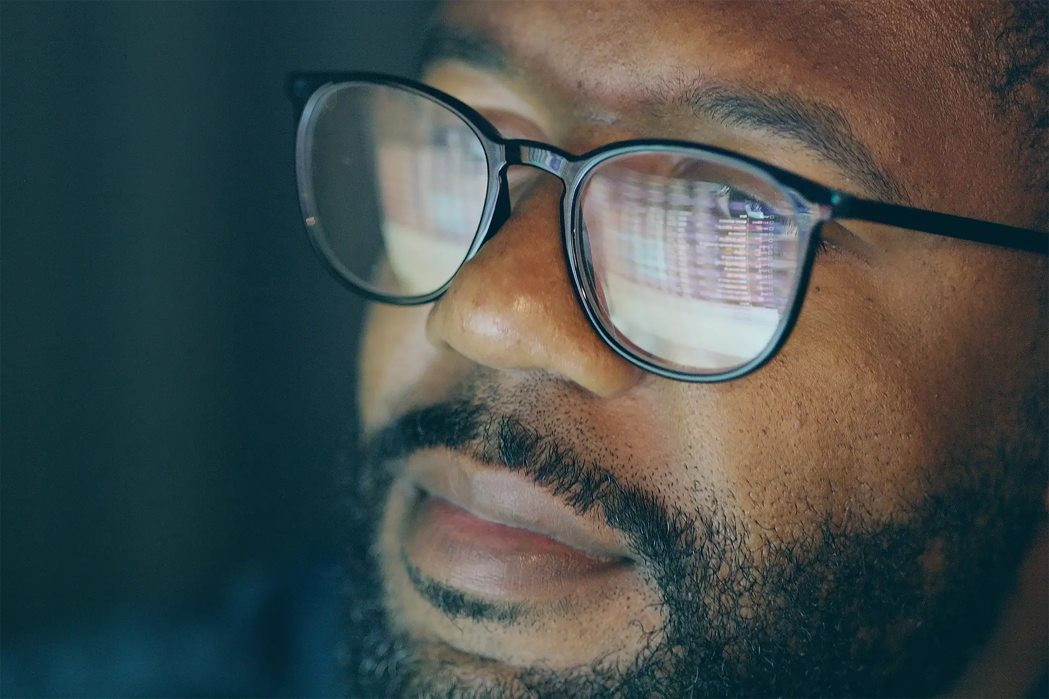 Close-up of a person with a beard and glasses, focusing intently on a computer screen. The screen's reflection is visible in the lenses, suggesting concentration or study. The lighting is dim, highlighting their face.