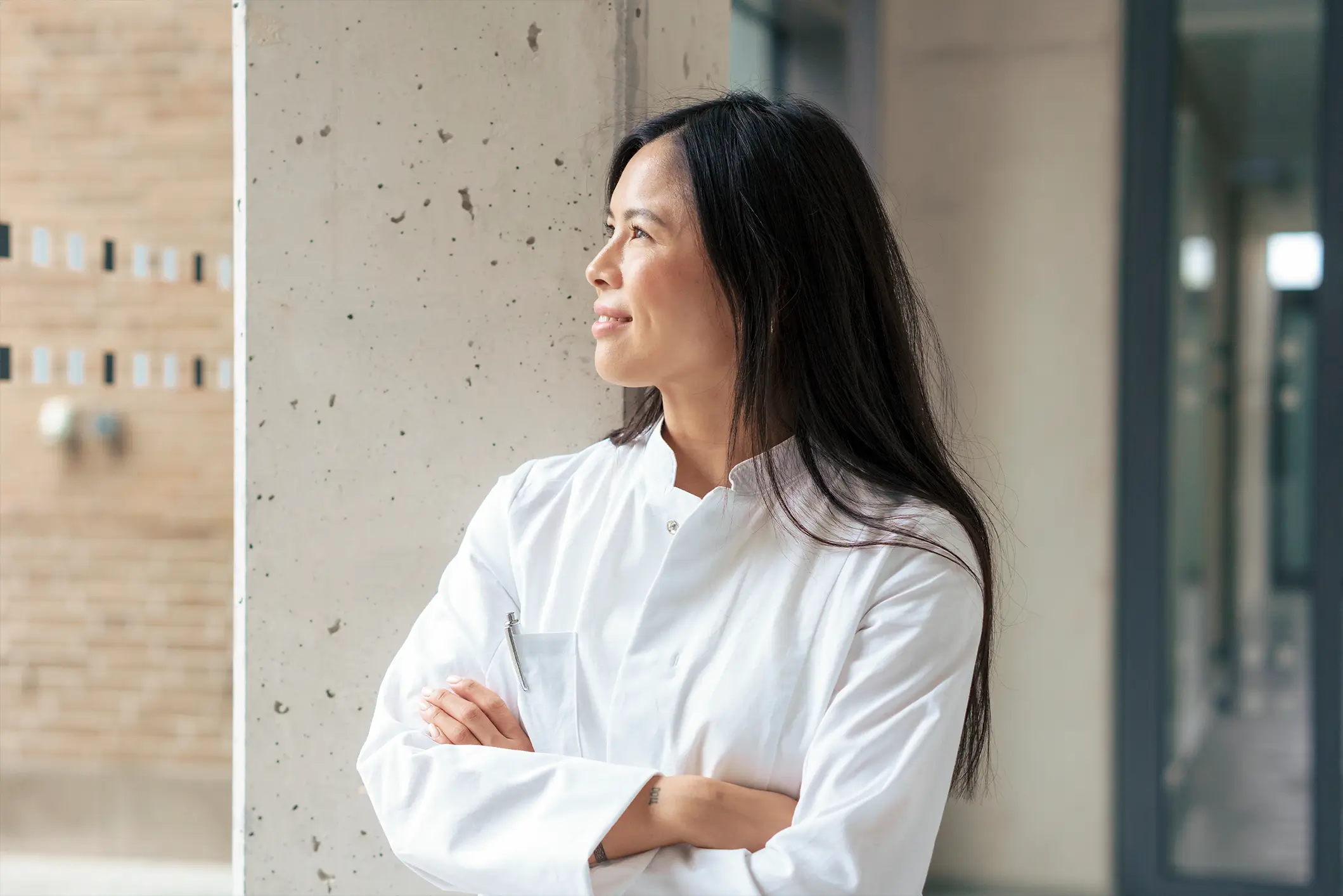 A woman wearing a white lab coat stands with her arms crossed, gazing thoughtfully to the side. She is in a brightly lit indoor space, with a textured concrete pillar and blurred hallway in the background.