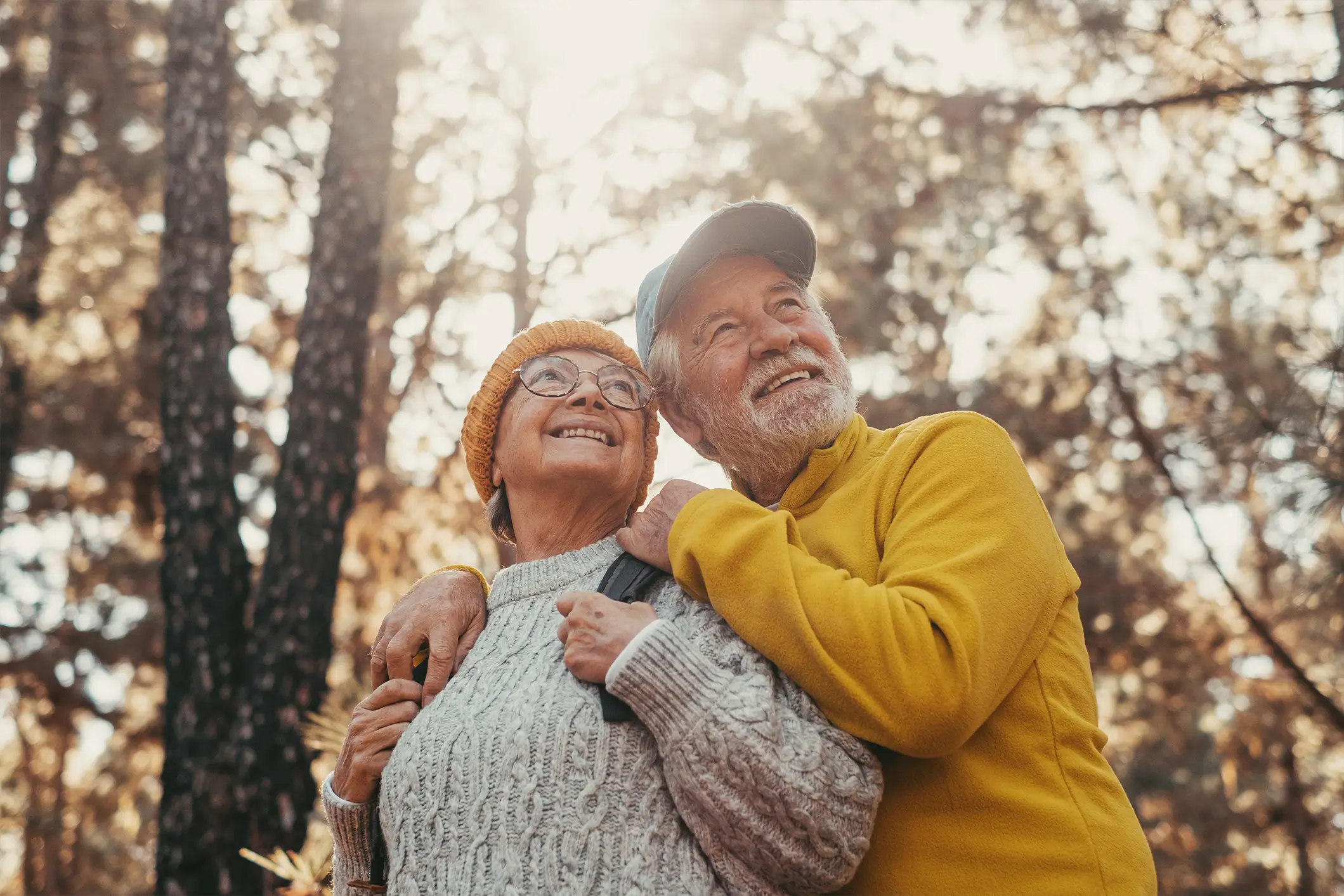 An elderly couple wearing warm clothing, with the man in a yellow jacket and blue cap, and the woman in a knit sweater and orange beanie, stand smiling and embracing each other in a sunlit forest with trees in the background.