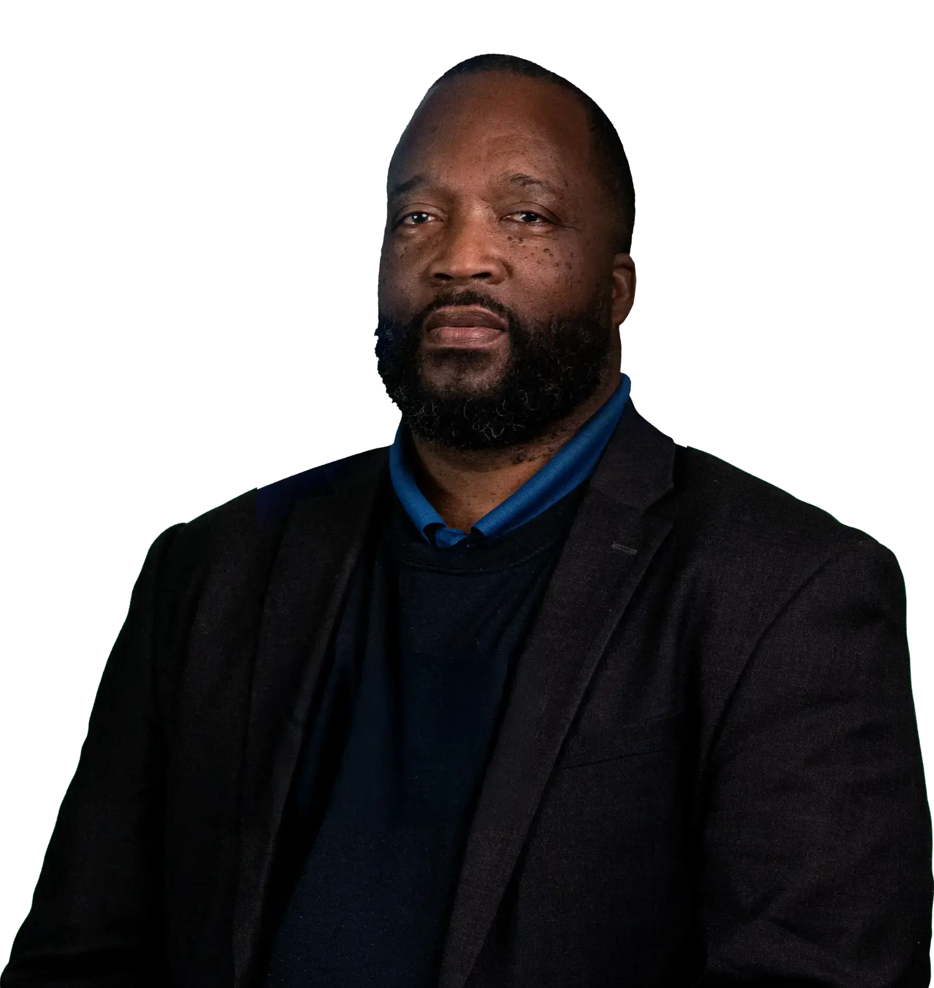 Man with a beard wearing a dark blazer and sweater, set against a black background, looking directly at the camera with a serious expression.