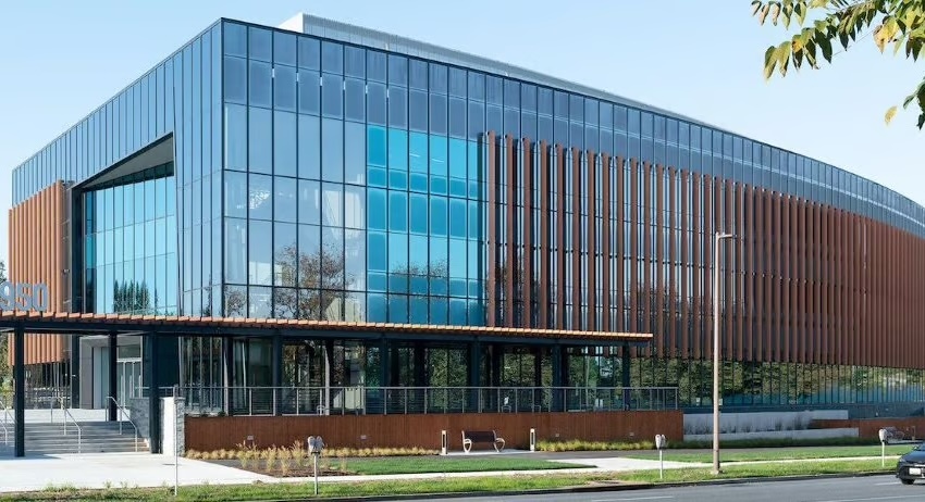 A modern building with large glass windows and vertical brown metal accents, surrounded by greenery and a sidewalk; the sky is clear and blue.