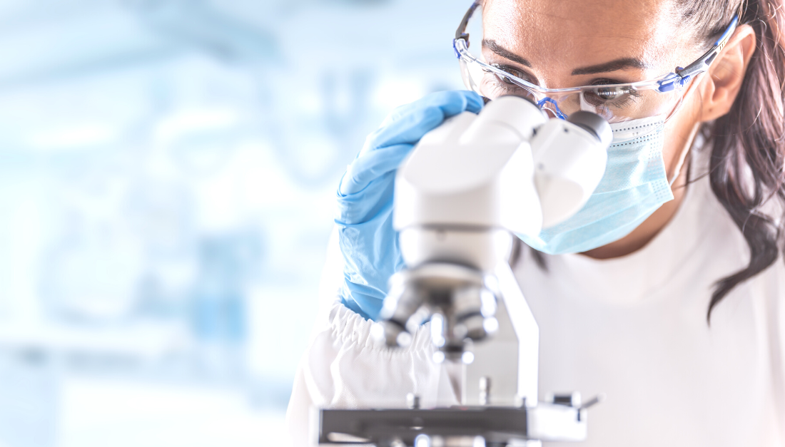 A scientist wearing safety goggles, a face mask, and blue gloves examines a sample through a microscope in a laboratory setting.