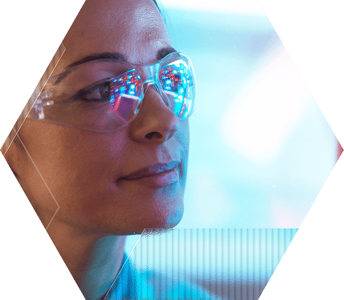 A person wearing safety glasses looks thoughtfully ahead, with colorful lights reflected in the lenses, set against a blue-toned background and framed in a hexagonal shape.