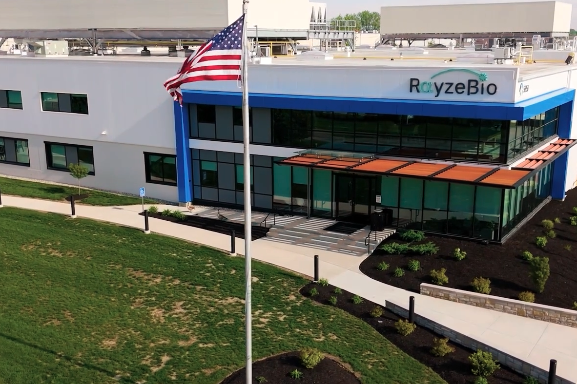 A modern building with a sign that reads "RayzeBio" above the entrance, large glass windows, landscaped grass and plants, and a U.S. flag flying on a pole in front of the building.
