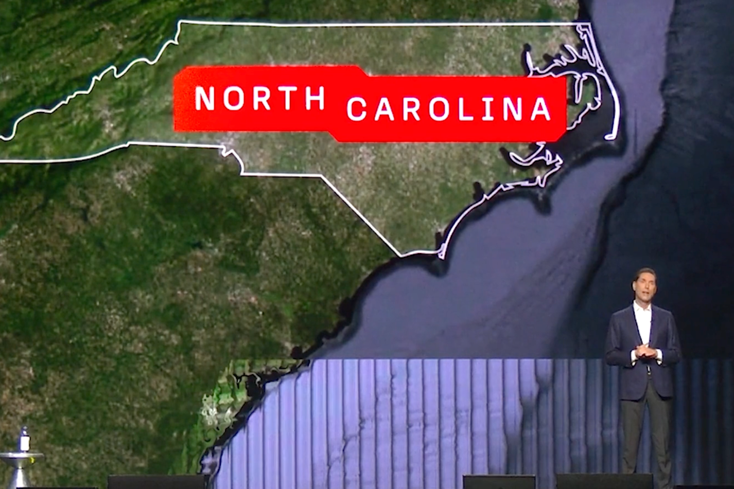 A person stands on stage beside a large screen displaying a map outline of North Carolina, labeled boldly in red with the words "NORTH CAROLINA.