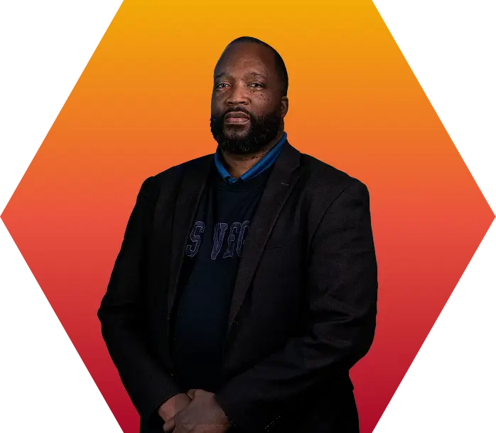 A man with a beard, wearing a dark blazer and shirt, stands against a gradient orange and red geometric background, looking forward with a neutral expression.