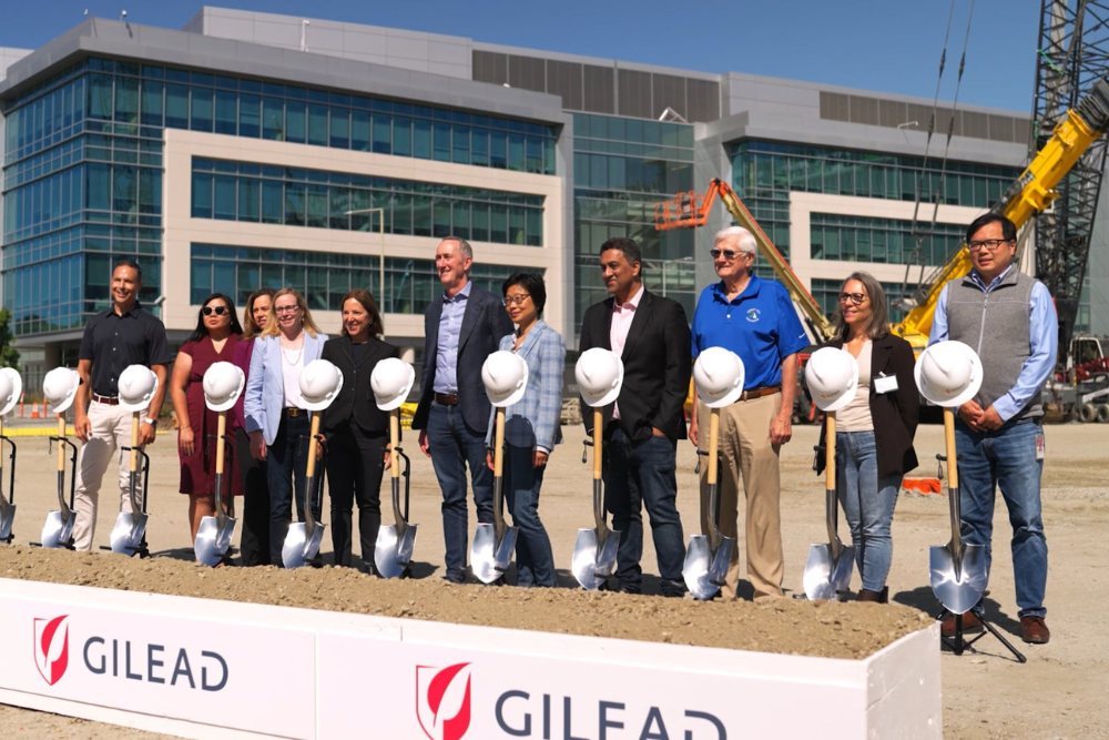 A group of people stands in front of a modern office building at a construction site, posing behind a row of shovels and white hard hats on a mound of dirt, with Gilead logos visible on the signs in front.