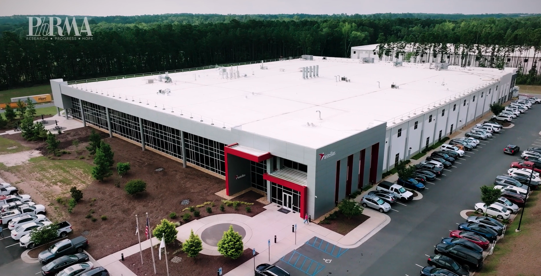 Aerial view of a large, modern industrial building with a white roof, surrounded by trees and a parking lot with cars. The entrance features red accents and a roundabout with landscaping.
