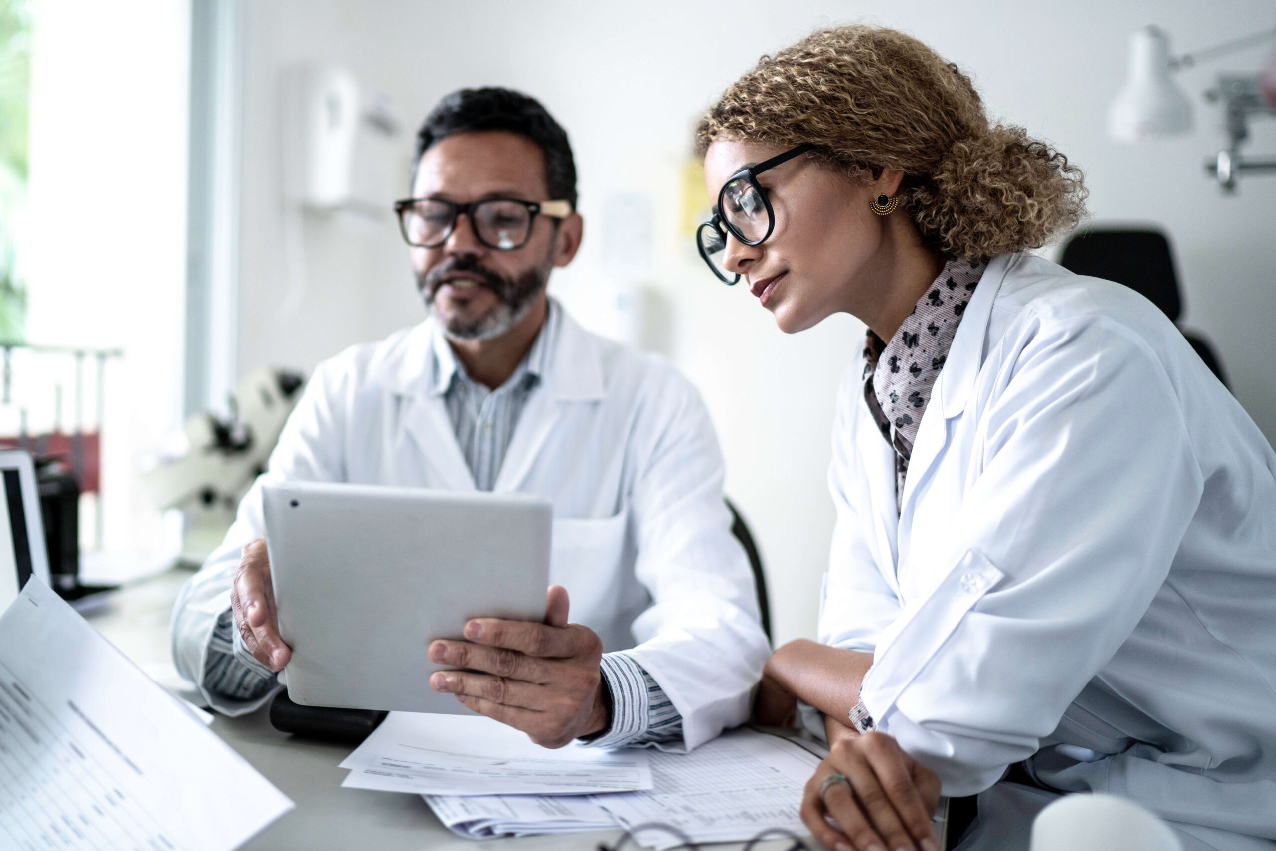 Two scientists wearing lab coats and glasses are sitting at a desk, looking at a tablet and reviewing documents together in a laboratory or office setting.