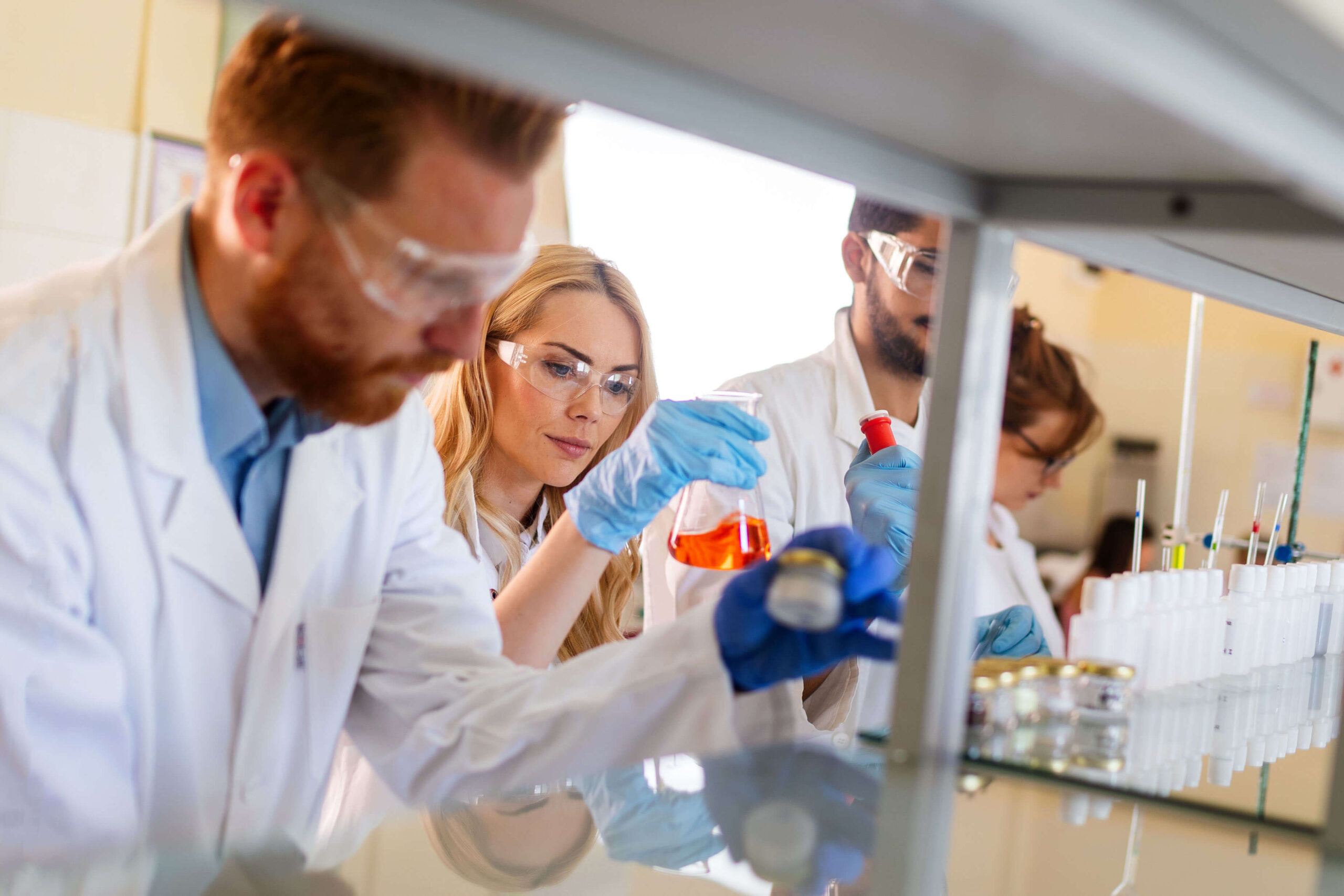 Three scientists wearing lab coats, gloves, and safety goggles work together in a laboratory, handling glassware and chemicals on a lab bench with test tubes and equipment in the background.