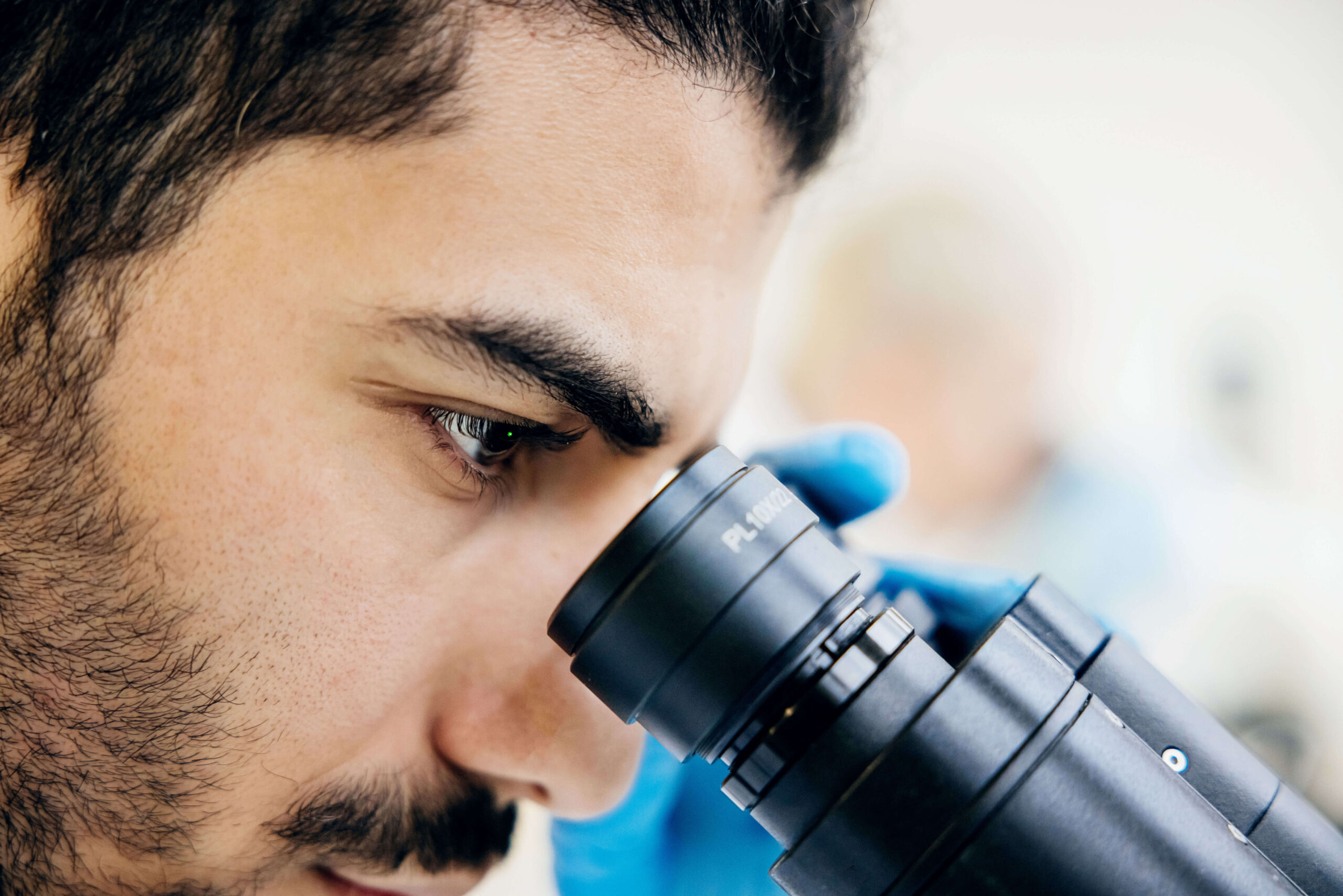 A close-up of a person with facial hair and blue gloves looking into a microscope, focusing intently on their work in a laboratory setting.