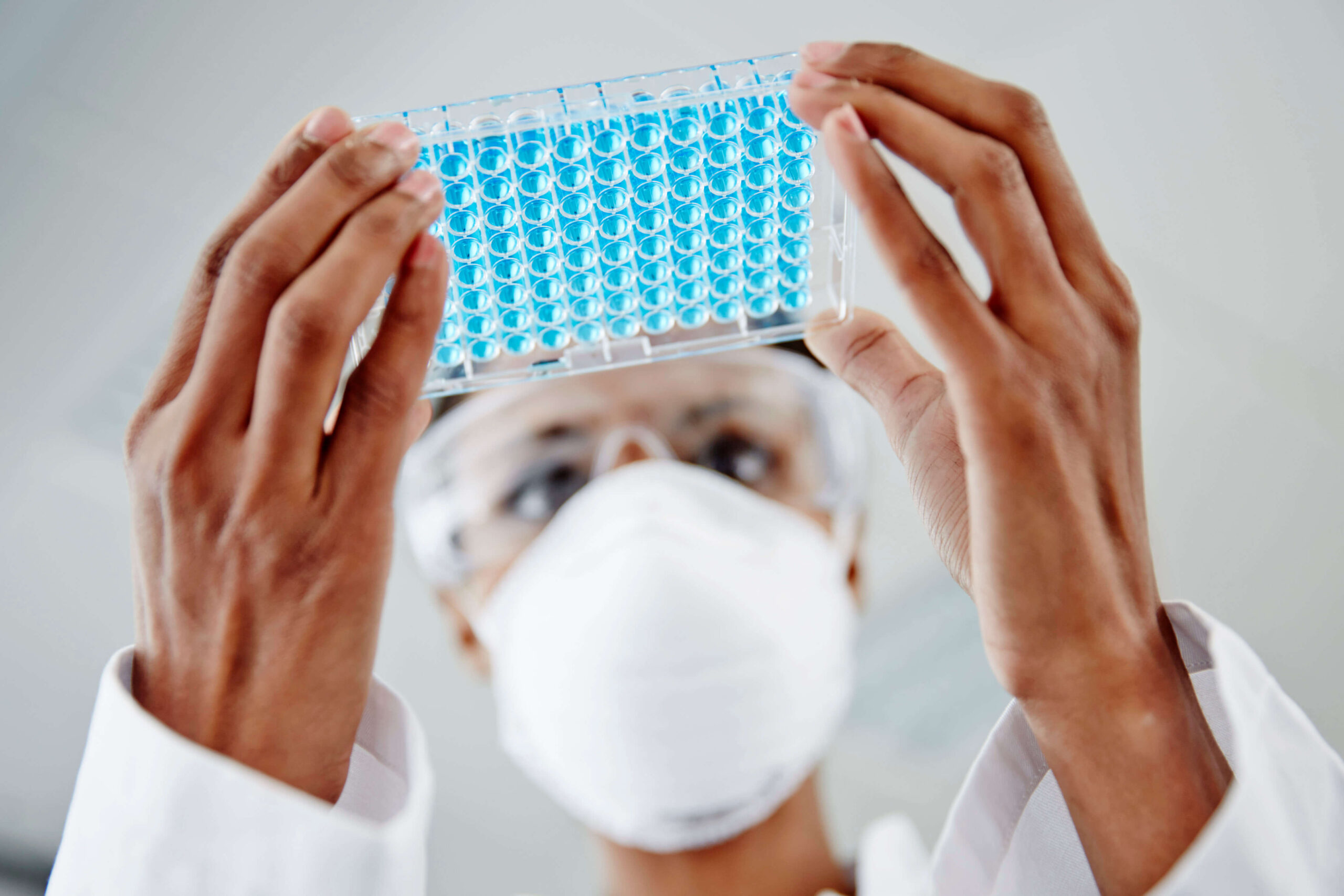 A scientist wearing safety goggles and a mask holds up a microplate filled with blue liquid, closely examining the samples in a laboratory setting.