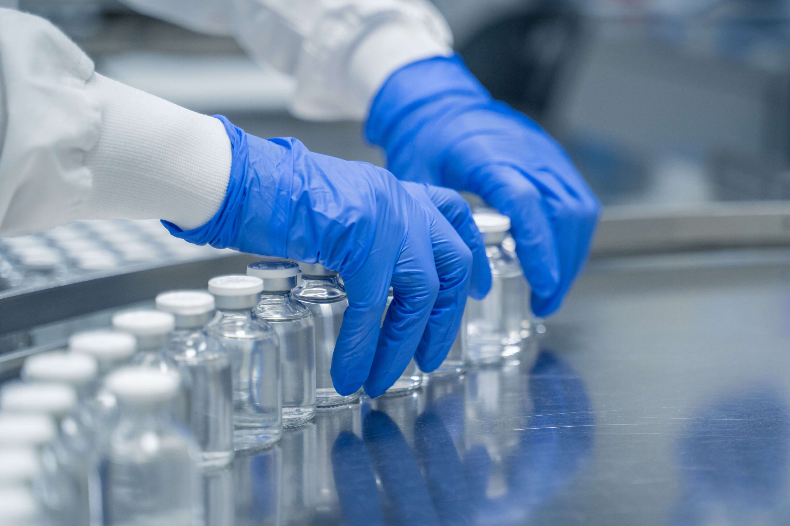 A person wearing blue gloves and a white lab coat is handling small glass vials filled with clear liquid, arranging them on a reflective metal surface in a laboratory setting.