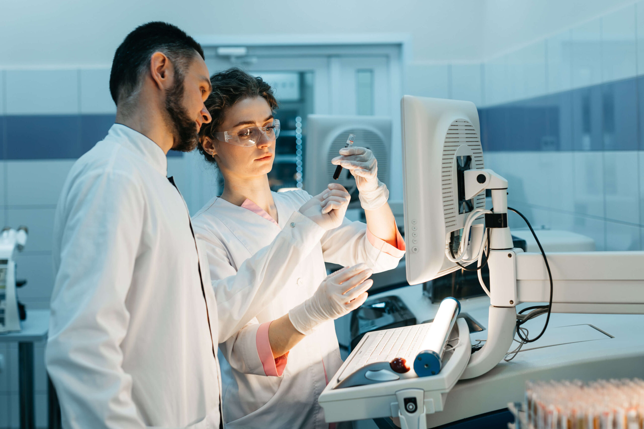 Two scientists in lab coats and gloves examine a vial together in a laboratory, standing next to a computer monitor and illuminated workspace with various lab equipment around them.