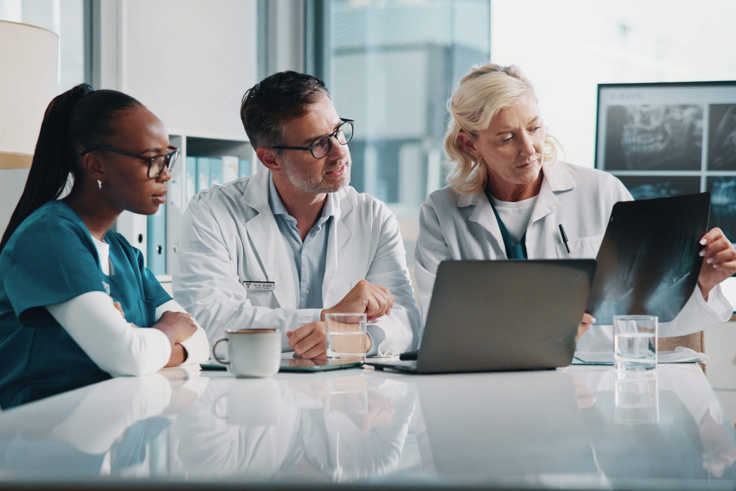 Three medical professionals sit at a table in an office, reviewing an X-ray together. A laptop, coffee mugs, and water glasses are on the table. Shelves and X-ray images are visible in the background.