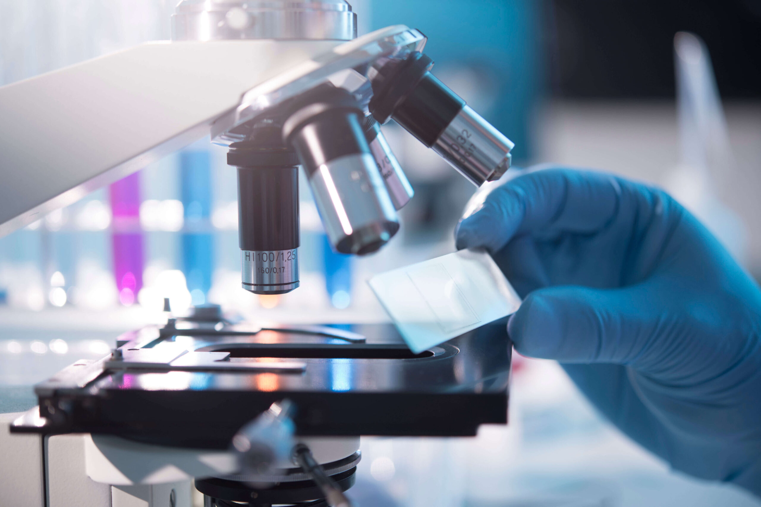 A gloved hand places a glass slide under the lens of a laboratory microscope, with blurred scientific equipment in the background.