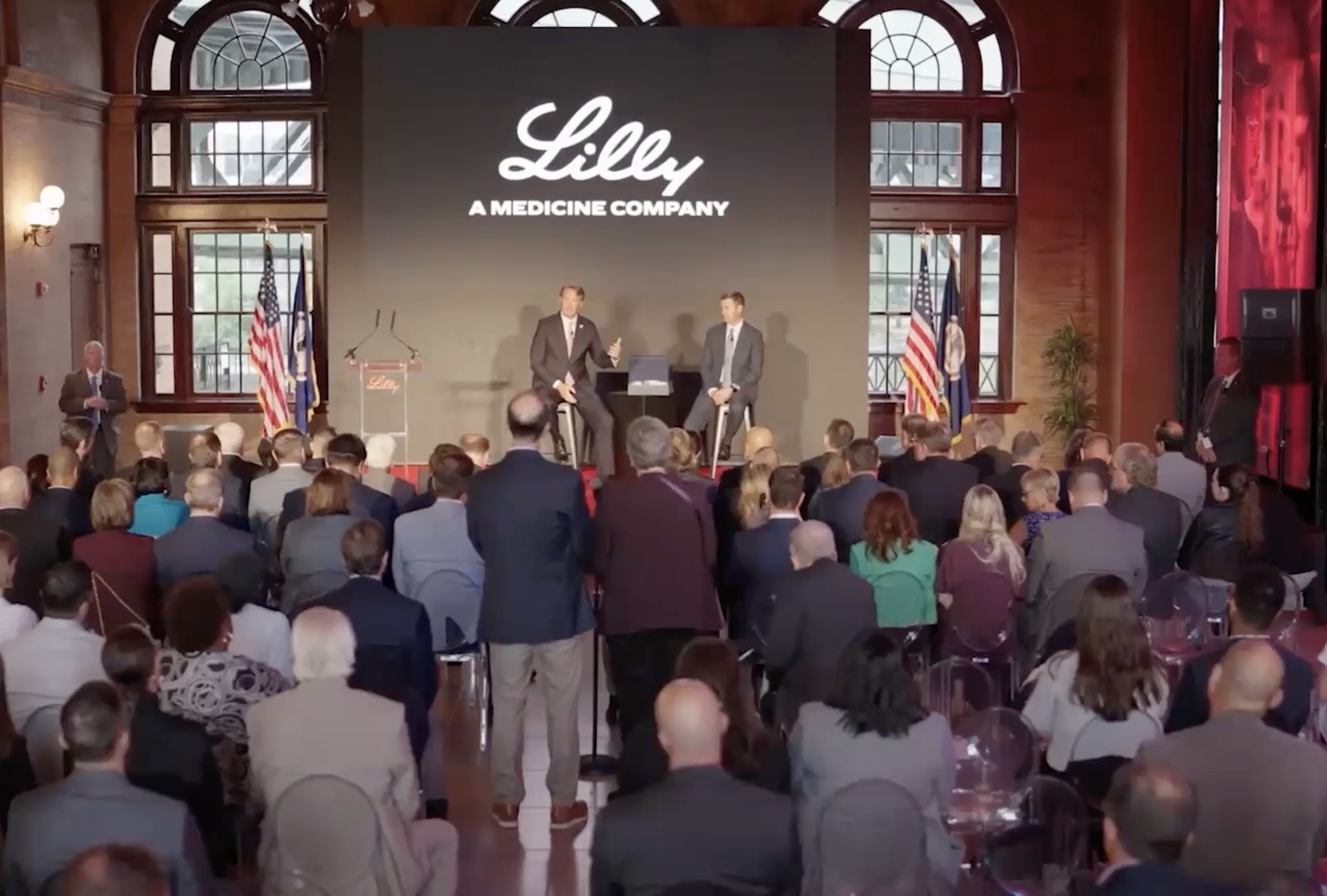 A large group of people sit facing a stage with two speakers, in front of a screen displaying the Lilly logo and “A Medicine Company,” in a room with tall windows and American flags.