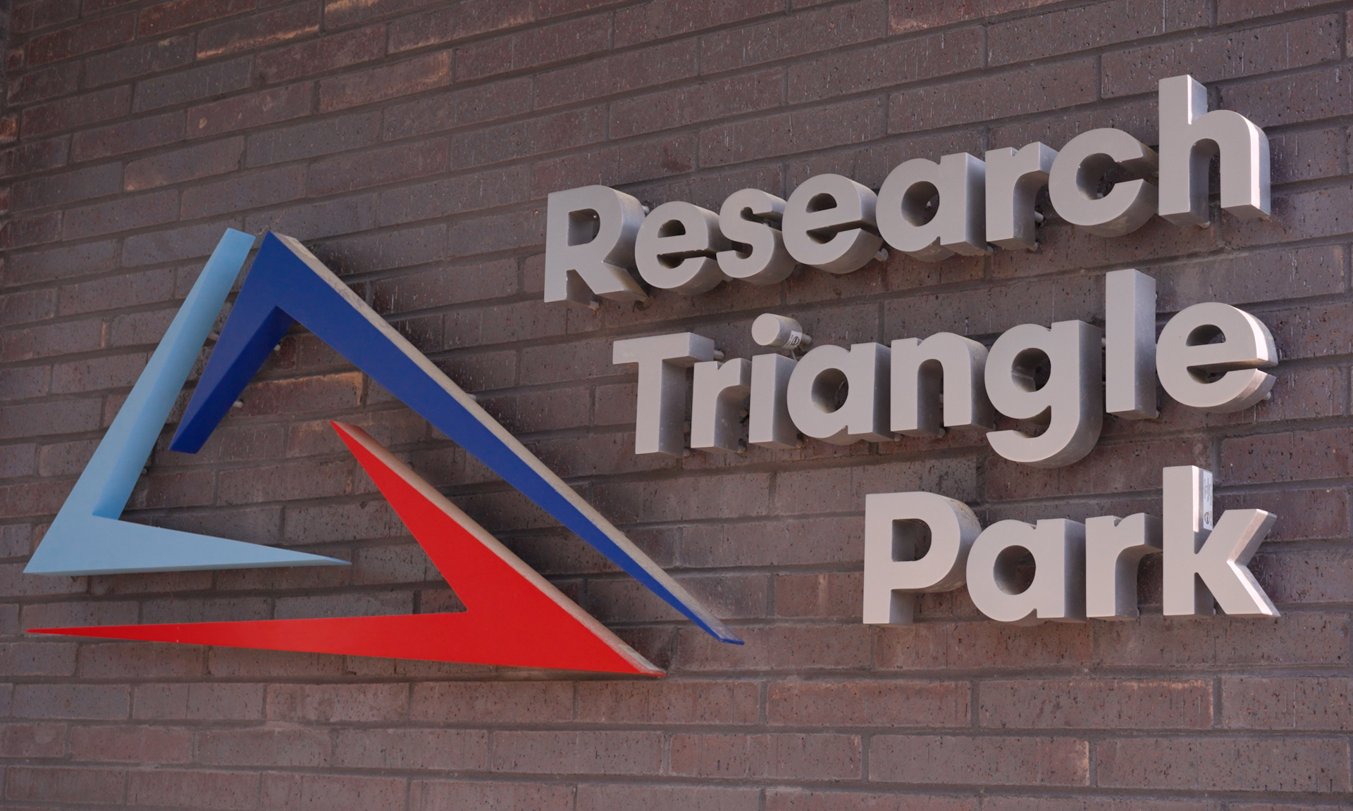 Silver 3D letters on a brick wall spell “Research Triangle Park” next to a geometric logo featuring blue and red angled shapes forming a triangle.