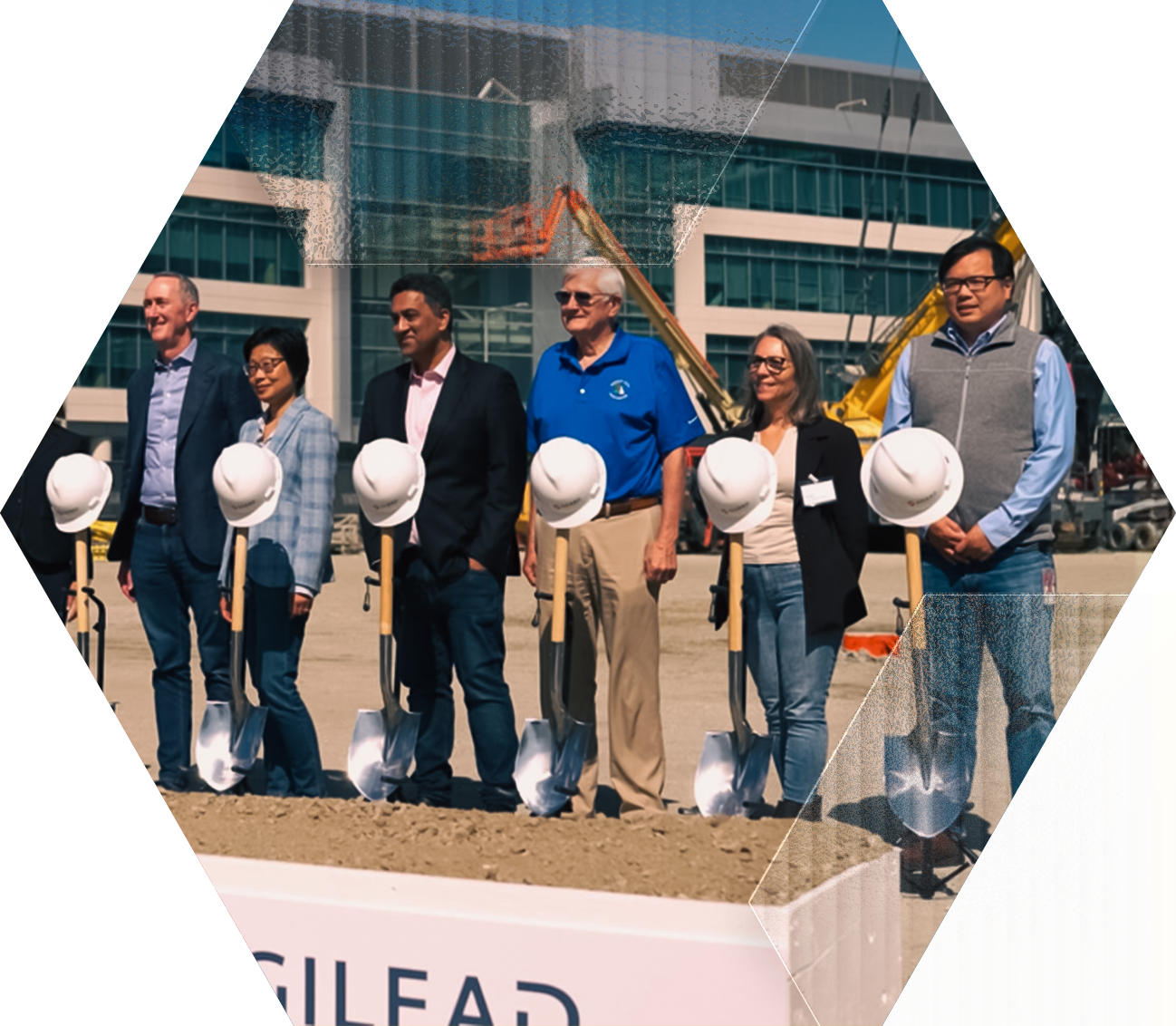 A group of seven people stand side by side outdoors, smiling, holding shovels at a groundbreaking ceremony. Helmets rest atop the shovels. A modern office building and construction equipment are in the background.