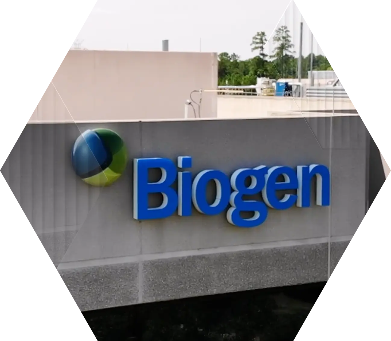 The image shows a building with a Biogen logo and sign in blue letters on a gray wall. The background includes trees and industrial rooftop structures.