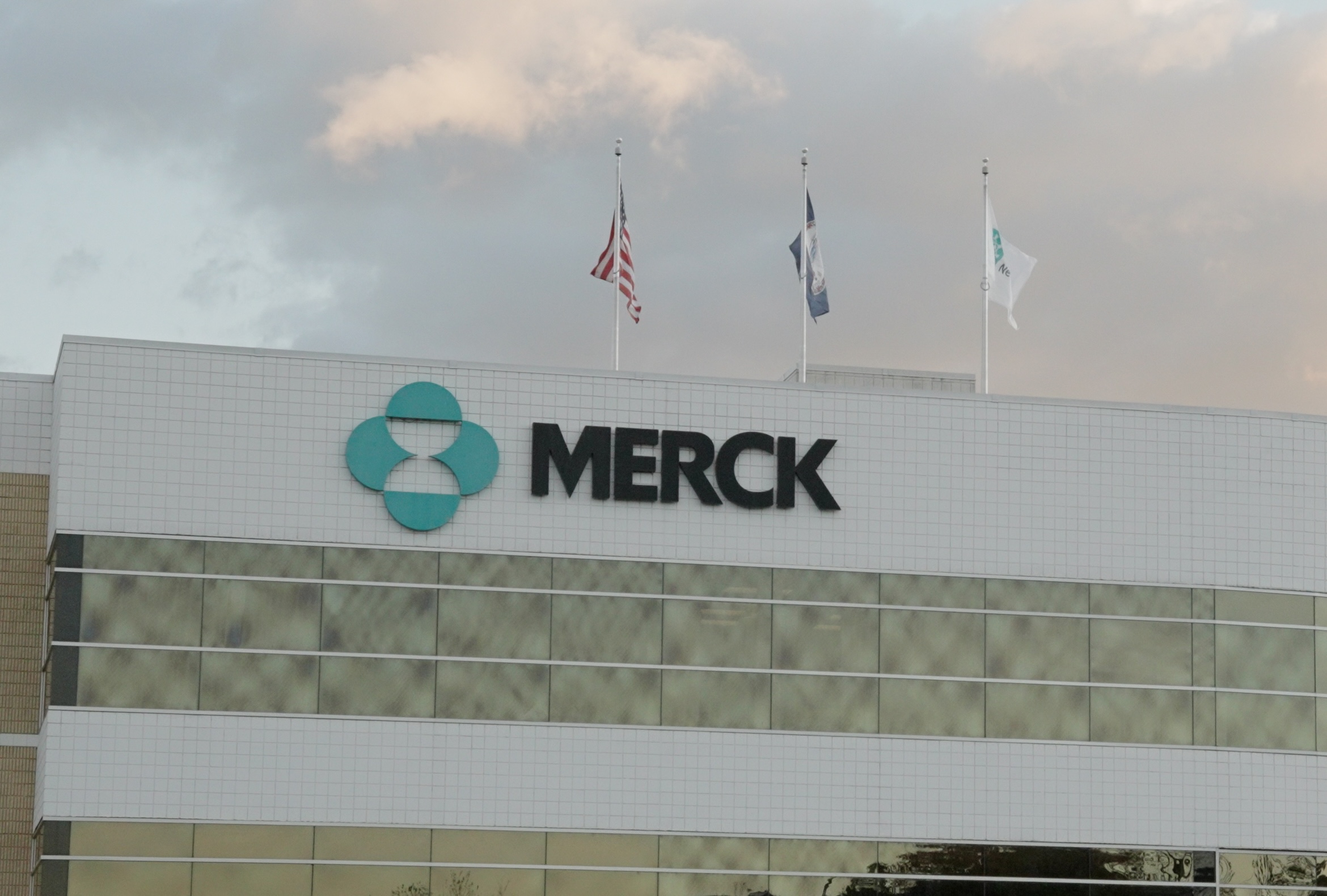 The image shows the exterior of a Merck building with the company’s logo and name on the facade. Three flags are flying on the roof against a cloudy sky.