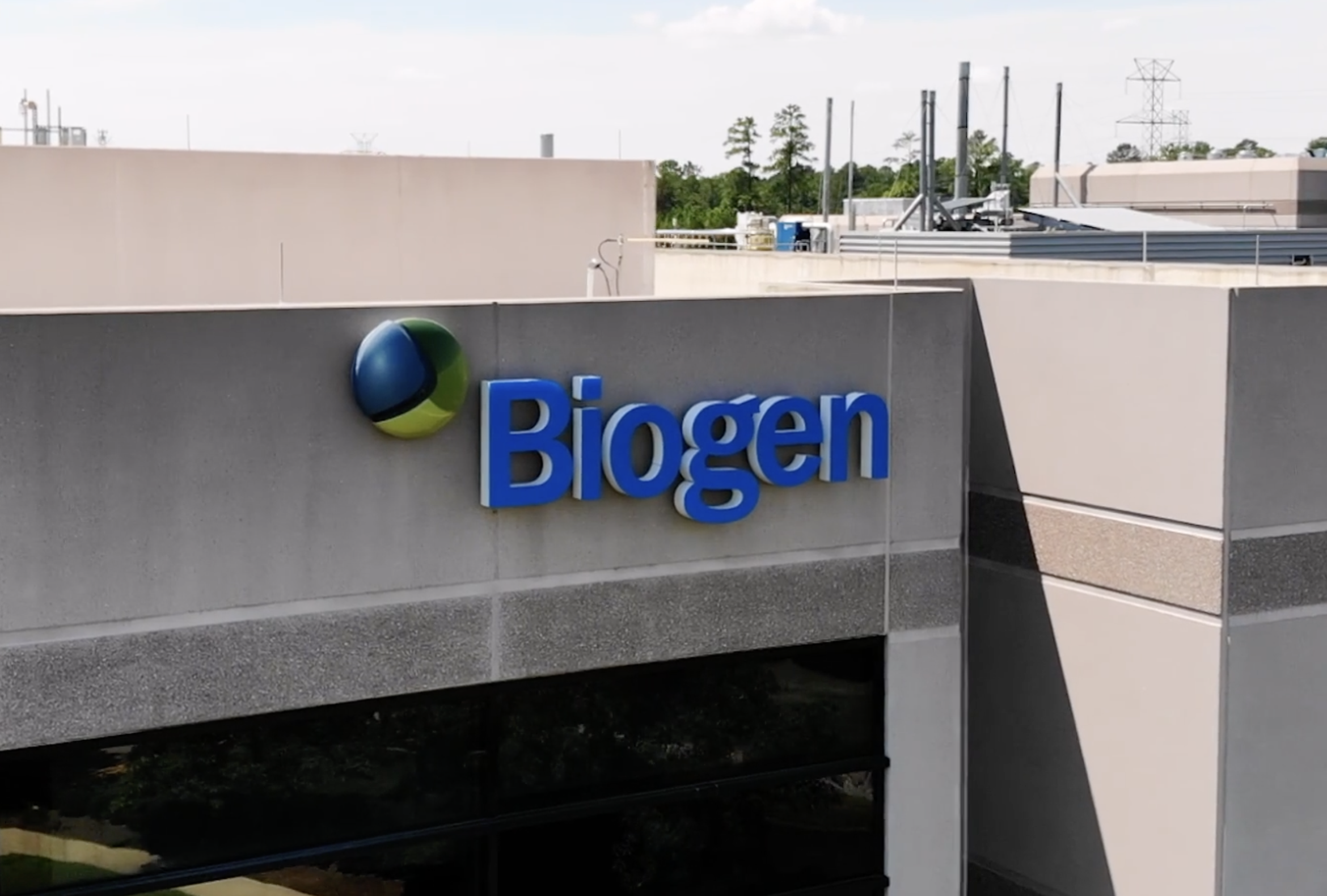 The exterior of a building with the Biogen logo and name displayed in blue letters on the wall; industrial equipment and trees are visible in the background.