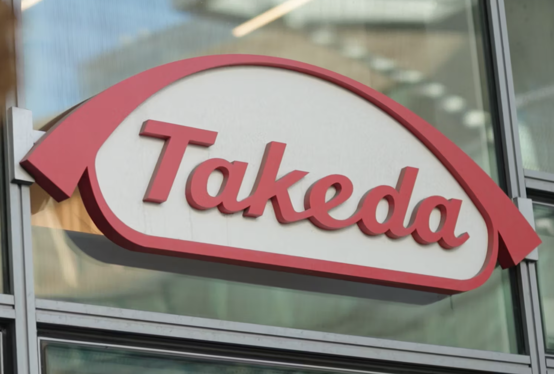 The image shows a Takeda logo sign in red and white on the exterior glass facade of a modern building.