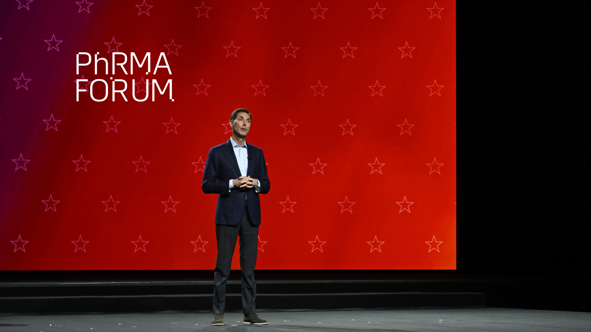 A person in a suit stands on stage in front of a red background with small stars and the words "PhRMA Forum" displayed on the left.
