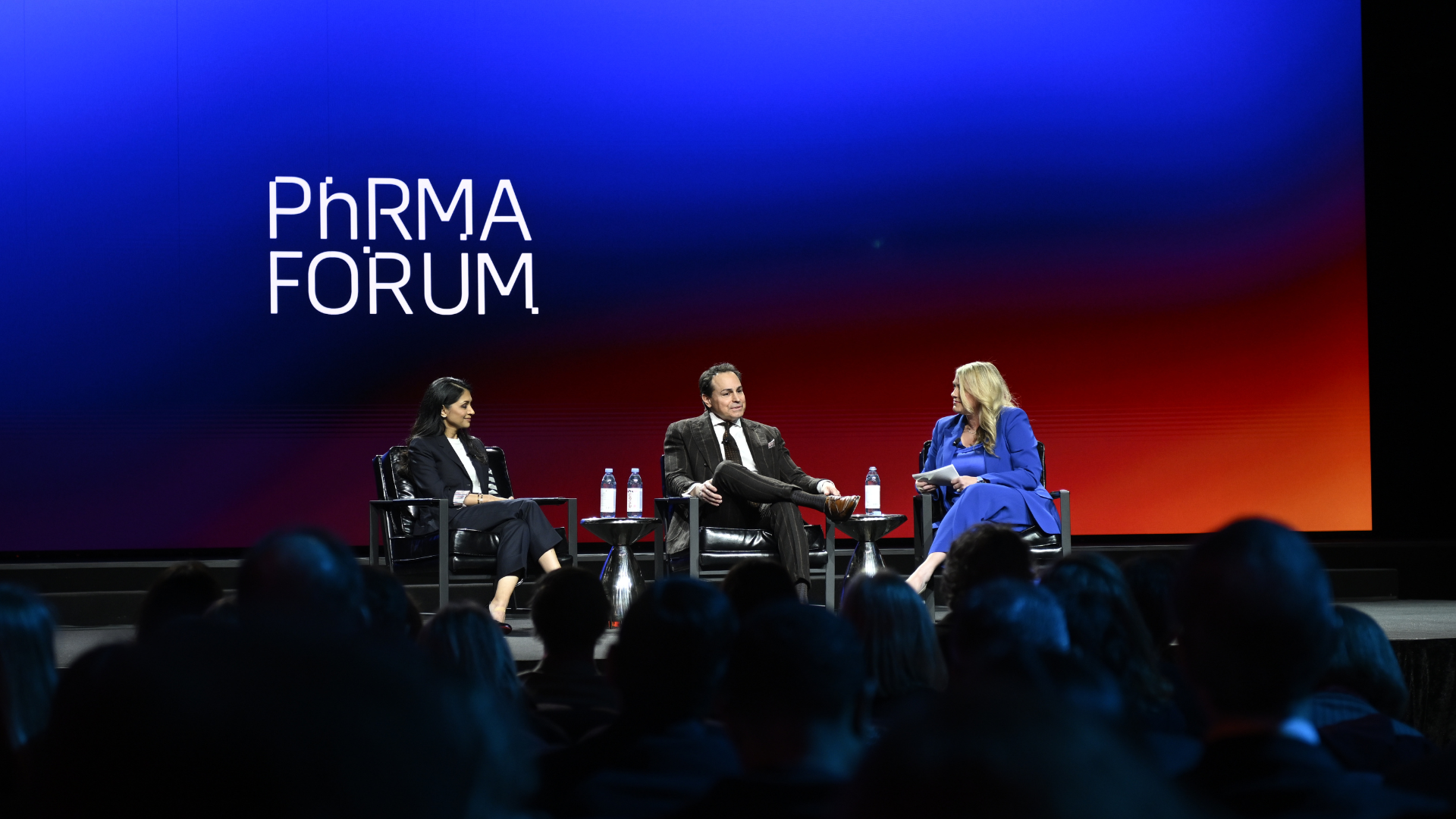 Three people sit onstage in armchairs, engaged in discussion at the PhRMA Forum. Two women and one man are visible, with an audience watching and a blue and red gradient backdrop displaying the event name.