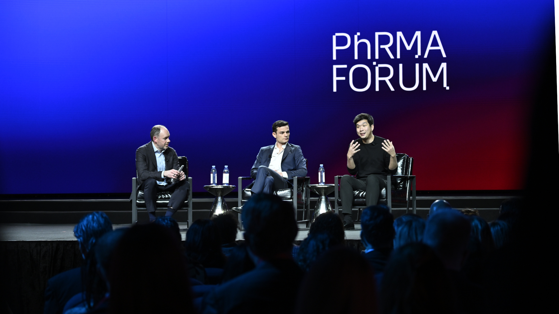 Three men sit on stage in armchairs, engaged in discussion at the PhRMA Forum. The background is a blue and red gradient with the event name displayed in large white letters. An audience watches in the foreground.