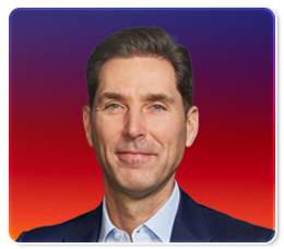 A man in a suit and light blue shirt smiles slightly in front of a gradient purple, red, and orange background.