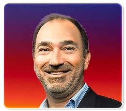 A middle-aged man with short dark hair and a beard, smiling, wearing a blue shirt and blazer. The background is a gradient of purple, red, and yellow tones.