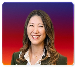 Smiling woman with long, wavy brown hair, wearing a white blouse and dark blazer, posed in front of a gradient background transitioning from purple to red to orange.