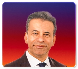 A man with short gray hair wearing a dark suit, white shirt, and dark tie, is smiling in front of a vibrant purple-to-red gradient background.