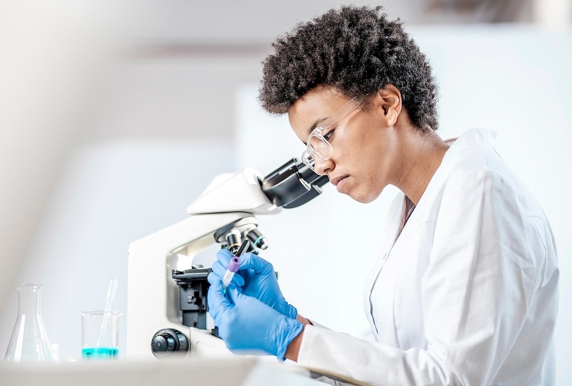 A scientist in a white lab coat and blue gloves examines a test tube while sitting at a microscope in a bright laboratory. Beakers with blue liquid are visible on the table.